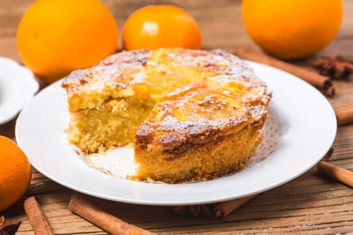 Fatia de um bolo de laranja com gengibre, decorado com fatias de laranja e açúcar de confeiteiro, em um prato branco.